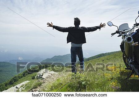 Man standing by motorcycle on top of mountain, arms outstretched View Large Photo Image Stock Image - Man standing by motorcycle on top of mountain, arms outstretched. Fotosearch