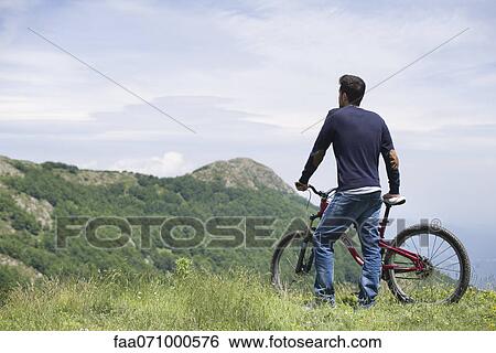 Man standing by mountain bike, enjoying scenic mountain view, rear view View Large Photo Image Stock Photograph - Man standing by mountain bike, enjoying scenic mountain view, rear view. Fotosearch