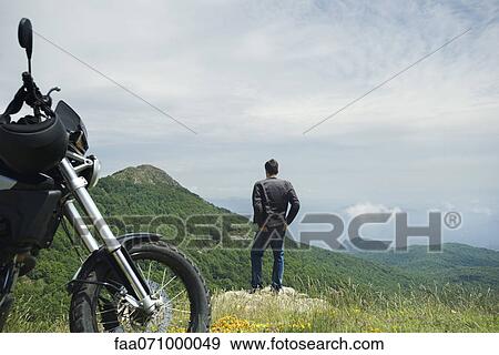 Man standing on rock looking at mountain view, motorcycle in foreground, rear view View Large Photo Image Stock Photo - Man standing on rock looking at mountain view, motorcycle in foreground, rear view. Fotosearch