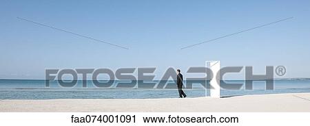 Stock Image - Businessman standing on beach next to half-open door, looking at ocean. Fotosearch