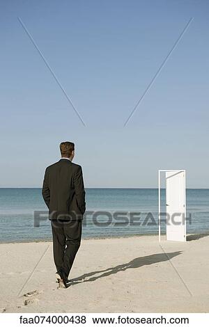 Stock Photo - Businessman walking on beach towards half-open door, rear view. Fotosearch