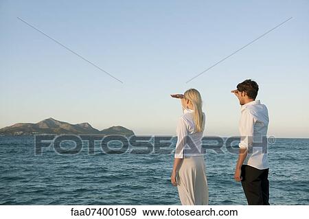 Couple standing by water looking at view View Large Photo Image Stock Photo - Couple standing by water looking at view. Fotosearch