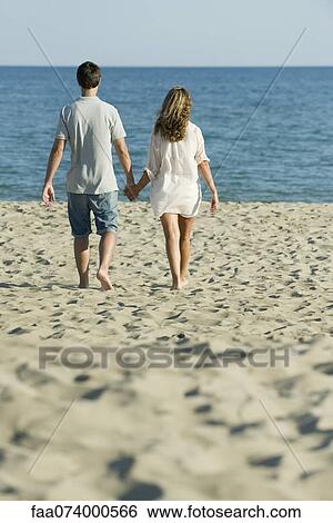Stock Photograph - Couple walking hand in hand at the beach, rear view. Fotosearch