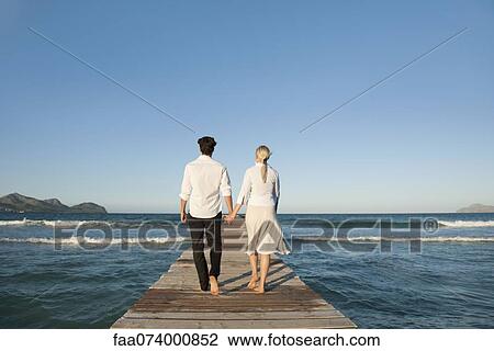 Stock Image - Couple walking on pier holding hands, rear view. Fotosearch