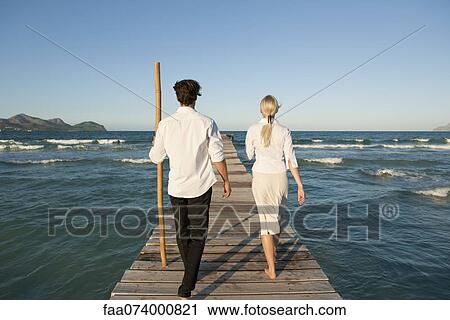 Stock Image - Couple walking on pier over water, rear view. Fotosearch