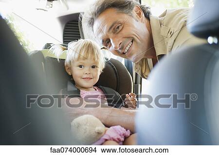 Picture - Father fastening little girl into car seat, both smiling at camera. Fotosearch