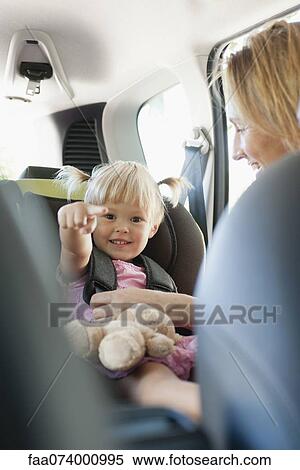 Stock Photography - Little girl pointing and smiling as her mother fastens her into car seat. Fotosearch