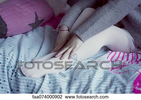 Stock Image - Little girl sitting on bed, touching feet, low section. Fotosearch