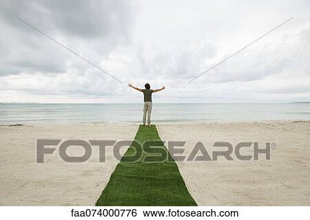 Stock Photograph - Man standing at end of carpet on beach with arms outstretched, rear view. Fotosearch