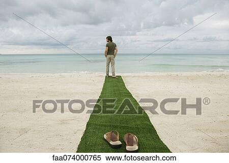Stock Photography - Man standing at end of carpet on beach, sandals on foreground. Fotosearch