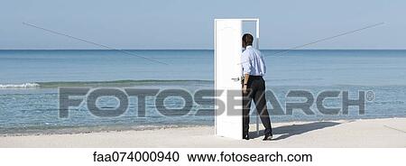 Stock Image - Man walking into open door on beach, rear view. Fotosearch