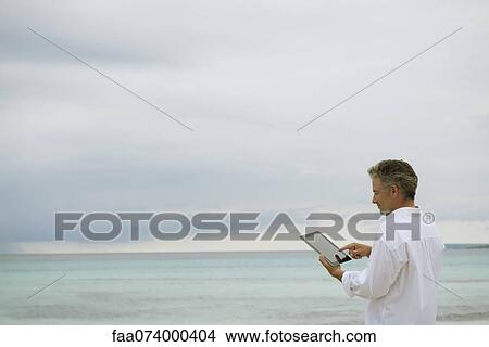 Picture - Mature man using digital tablet by ocean. Fotosearch