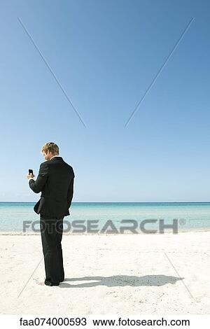 Young businessman using cell phone while standing on beach View Large Photo Image Stock Image - Young businessman using cell phone while standing on beach. Fotosearch