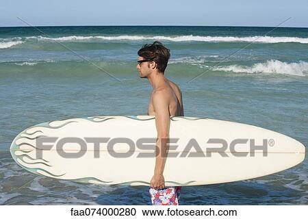 Stock Image - Young man carrying surfboard, side view. Fotosearch
