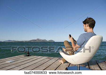 Young man sitting in armchair by lake using cell phone, side view View Large Photo Image Stock Image - Young man sitting in armchair by lake using cell phone, side view. Fotosearch