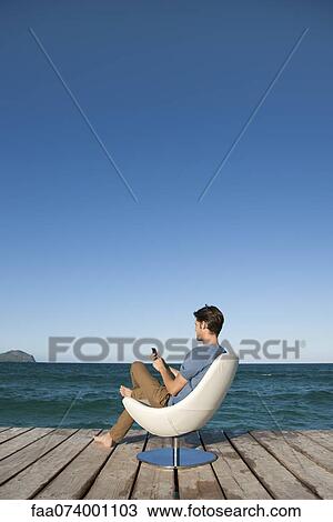 Young man sitting in armchair by lake using cell phone, side view View Large Photo Image Stock Image - Young man sitting in armchair by lake using cell phone, side view. Fotosearch