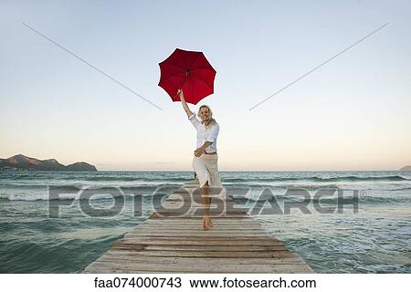 Stock Image - Young woman jumping on pier holding umbrella. Fotosearch