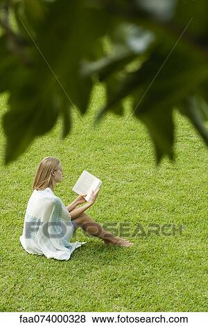 Stock Photo - Young woman sitting on grass reading book, high angle view. Fotosearch