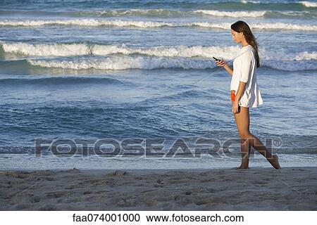 Stock Image - Young woman using cell phone while strolling on beach, side view. Fotosearch
