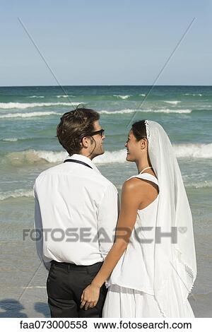 Stock Photo - Bride and groom standing on beach, looking at view. Fotosearch