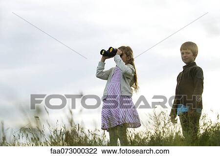 Stock Image - Children standing in field, girl looking through binoculars. Fotosearch