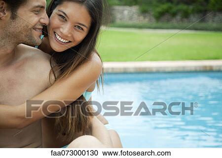 Stock Image - Couple embracing by swimming pool, portrait. Fotosearch