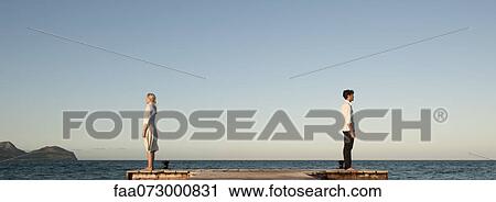 Stock Image - Couple standing back to back on pier, side view. Fotosearch