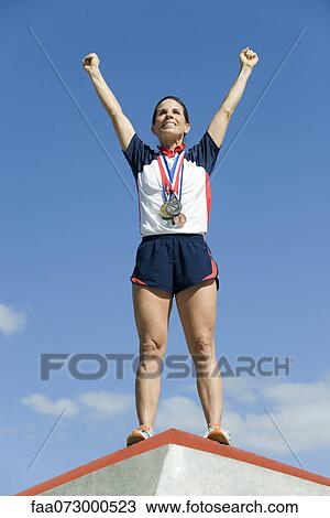 Female athlete standing on winner's podium with arms raised in victory View Large Photo Image Stock Image - Female athlete standing on winner's podium with arms raised in victory. Fotosearch