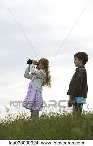 Picture - Girl standing in field with boy, looking through binoculars. Fotosearch
