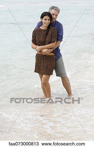 Stock Image - Husband and wife embracing at beach, ankles deep in water. portrait. Fotosearch