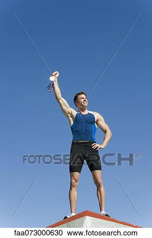 Stock Image - Male athlete on podium, holding up gold medal. Fotosearch