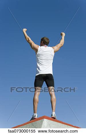 Male athlete standing on winner's podium with arms raised in victory, rear view View Large Photo Image Stock Photography - Male athlete standing on winner's podium with arms raised in victory, rear view. Fotosearch