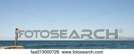 Man in distance standing on pier looking at ocean view through binoculars View Large Photo Image Stock Photograph - Man in distance standing on pier looking at ocean view through binoculars. Fotosearch