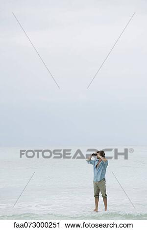 Stock Image - Man standing in sea, looking through binoculars. Fotosearch