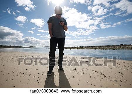 Stock Image - Man standing on beach, looking at cell phone. Fotosearch