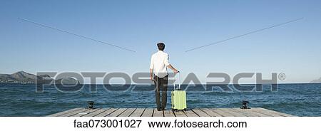 Stock Photo - Man standing on dock with suitcase, looking at ocean view, rear view. Fotosearch