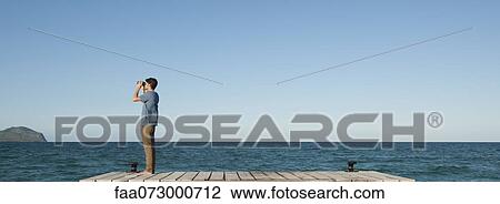 Stock Image - Man standing on pier looking at ocean view through binoculars, side view. Fotosearch