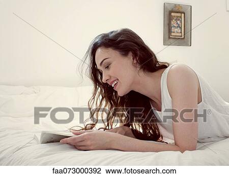 Stock Image - Mid-adult woman reading book on bed. Fotosearch