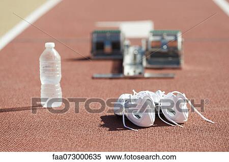 Stock Photography - Running shoes and water bottle on running track, starting blocks in background. Fotosearch