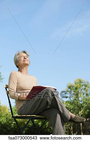 Stock Image - Senior woman sitting outdoors with book, enjoying warmth of sun. Fotosearch