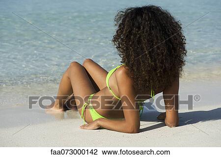 Stock Image - Woman relaxing on beach, looking at ocean, rear view. Fotosearch