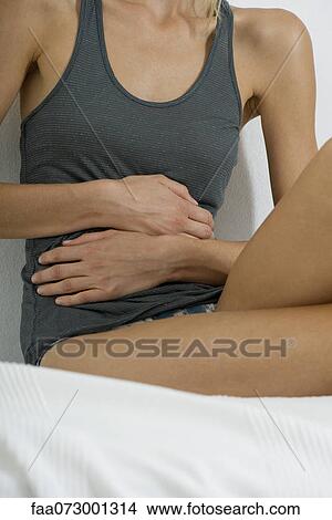 Picture - Woman sitting on bed, hands on stomach, mid section. Fotosearch
