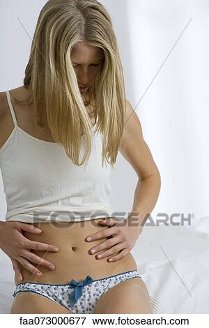 Woman sitting on bed, looking down at abdomen View Large Photo Image Stock Photo - Woman sitting on bed, looking down at abdomen. Fotosearch