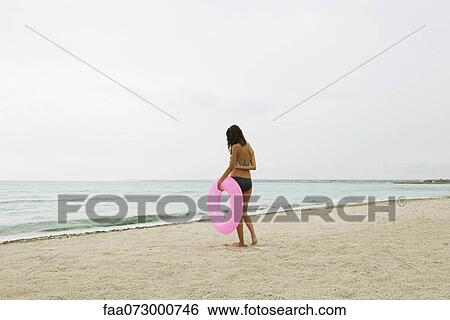 Stock Photograph - Woman standing on beach, holding inflatable ring, rear view. Fotosearch