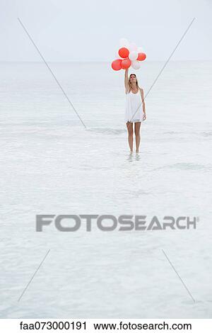 Stock Image - Woman standing on surface of water, holding bunch of balloons. Fotosearch