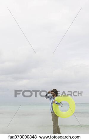 Young man carrying inflatable swimming ring, looking into distance View Large Photo Image Stock Photo - Young man carrying inflatable swimming ring, looking into distance. Fotosearch