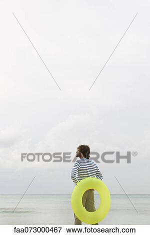Stock Photo - Young man holding inflatable ring, looking at ocean view. Fotosearch