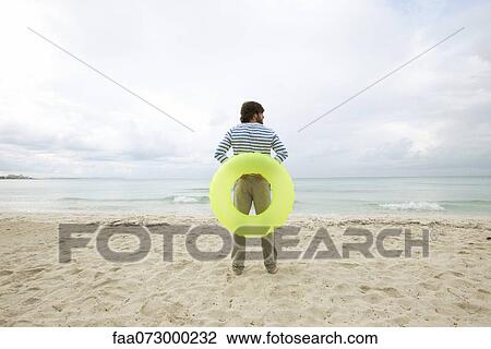 Young man staning on beach with inflatable ring, rear view View Large Photo Image Stock Image - Young man staning on beach with inflatable ring, rear view. Fotosearch