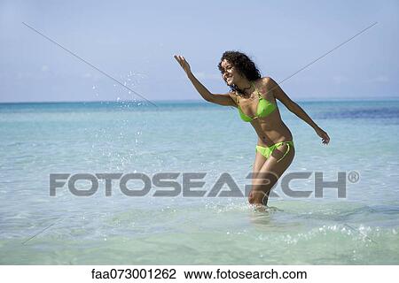 Stock Image - Young woman in bikini splashing water in ocean. Fotosearch