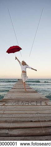Stock Image - Young woman jumping on pier holding umbrella. Fotosearch
